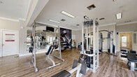 Located at 100 Arbor Lake Boulevard in Hermitage, Tennessee, this fitness center offers a generously sized, single-floor exercise room designed to support a full range of workout routines. The space features durable wood-look flooring throughout, crown molding, and bright flush-mounted lighting that creates a clean, energizing environment. Large windows and glass-paned doors allow natural light to flow in while providing views of the outdoors, contributing to an open and motivating atmosphere. Full-length mirrors line portions of the walls, enhancing the sense of space and allowing users to monitor their form during workouts. The exercise room is comprehensively equipped with both cardio and strength training machines, including treadmills, stationary bikes, ellipticals, cable machines, Smith machines, weight benches, and free weights. Stability balls and medicine balls are also available for functional training. Wall-mounted televisions are positioned throughout the room for entertainment during workouts. A bold accent wall featuring an inspirational graphic reading "energy. fit. strength." adds a dynamic visual focal point to one section of the room, reinforcing the fitness-forward character of the space. The layout is thoughtfully organized to provide ample room to move between equipment safely and comfortably.