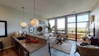 This single-floor, two-bedroom, two-bathroom residence in Sheboygan, Wisconsin offers a thoughtfully designed open-concept layout with modern finishes throughout. The living area is anchored by large floor-to-ceiling windows that flood the space with natural light and frame expansive views of the surrounding cityscape. A sliding glass door opens directly onto a private balcony, extending the living space outdoors.

The kitchen is well-appointed with dark espresso cabinetry, granite countertops, a mosaic tile backsplash, and stainless steel appliances including a refrigerator, range, and built-in microwave. A granite-topped island with bar seating serves as a natural divider between the kitchen and living area, with pendant lighting overhead adding a stylish touch. Wood-style flooring runs throughout the main living spaces.

Both bedrooms are carpeted and feature large windows with open views. Each bedroom is served by its own full bathroom, finished with dark vanity cabinetry, granite countertops, tile flooring, and glass-enclosed walk-in showers. The overall ambiance is contemporary and bright, combining clean architectural lines with warm, inviting finishes.