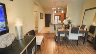 This well-appointed single-floor space offers a thoughtfully designed open-concept layout that connects the living, dining, and kitchen areas into one cohesive and functional environment. Rich hardwood flooring flows throughout, lending warmth and continuity to the space.

The kitchen is a standout feature, showcasing warm-toned cabinetry, stainless steel appliances, and a breakfast bar peninsula that doubles as a natural divider between the kitchen and dining area. Pendant lighting with a modern track-style fixture illuminates the kitchen, adding both function and visual interest.

The dining area comfortably accommodates a full-sized table and seating, making it ideal for everyday meals or entertaining. A hallway extends from the main living area, leading to additional rooms beyond, including what appears to be a bathroom with a classic door detail. The entryway features a bold, dark front door that creates a striking contrast against the light-toned walls.

Throughout the space, neutral wall colors paired with the warm flooring and contemporary furnishings create a balanced, inviting ambiance. The layout is efficient yet spacious-feeling, with clear sightlines across the main living areas that contribute to an open and airy atmosphere.