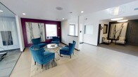 This single-floor space makes a strong first impression with its thoughtfully designed interior that balances modern aesthetics with practical functionality. The lounge area serves as a focal point, anchored by a set of rich teal velvet accent chairs arranged around a sleek glass coffee table atop a light-toned area rug. A bold plum-colored accent wall adds depth and visual drama, contrasting beautifully against the clean white walls and light-colored flooring that runs throughout the space.

Natural light flows in through multiple windows, brightening the open layout and enhancing the airy atmosphere. Recessed ceiling lighting supplements the natural light, ensuring the space remains well-illuminated at all times. A large abstract artwork adorns the accent wall, adding a sophisticated artistic touch to the lounge setting.

Beyond the lounge, the layout opens into additional areas, including what appears to be a reception or administrative zone, complete with a glass partition and directory display. The thoughtful use of space, clean lines, and carefully curated furnishings give this interior a polished, professional character, making it well-suited for welcoming guests or facilitating day-to-day operations in a stylish and organized environment.