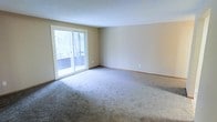 This single-floor space centers around a spacious living area finished with wall-to-wall carpet in a neutral gray-beige tone, providing a comfortable and understated foundation throughout the room. The walls are painted in a soft, warm beige, contributing to a clean and open atmosphere. A large sliding glass door serves as a prominent architectural feature, allowing an abundance of natural light to fill the room while providing direct access to an outdoor balcony or patio area. Through the glass, views of mature trees and natural greenery are visible, adding a sense of connection to the outdoors. The room's layout is open and unobstructed, offering flexible arrangement possibilities. Neutral-toned baseboards frame the perimeter of the room, complementing the overall color palette. The flat, textured ceiling and straightforward design keep the aesthetic simple and functional. The combination of natural light, soft carpeting, and outdoor access creates a calm, livable environment well-suited for everyday use.