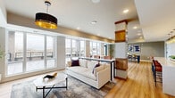 Located at 9900 56th Ave N in Plymouth, Minnesota, this thoughtfully designed single-floor space seamlessly integrates a living room, kitchen, and dining room into one cohesive open-concept layout. Light hardwood flooring flows throughout, unifying the distinct functional areas beneath recessed lighting and designer pendant fixtures.

The living room anchors the space with a striking stone-clad fireplace paired with a wall-mounted television, accented by a bold navy feature wall and vertical wood slat detailing. A dramatic drum pendant light adds a sophisticated touch. Large floor-to-ceiling windows and a glass door open to a spacious outdoor terrace, flooding the interior with natural light and offering expansive outdoor views.

The kitchen is a standout feature, showcasing a generous quartz-topped island with leather barstool seating, white cabinetry with brushed gold hardware, and a coordinated series of globe pendant lights suspended above. A navy accent soffit and open shelving add character and depth to the design.

The dining area comfortably accommodates a full dining table beneath elegant gold cluster pendants, with wraparound windows providing additional natural light and terrace access. Structural columns with warm wood-capped detailing serve as architectural accents, adding visual interest throughout this refined and well-appointed space.
