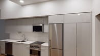 This single-floor space showcases a thoughtfully designed, contemporary interior with a cohesive minimalist aesthetic. The kitchen is a standout feature, outfitted with flat-front gray cabinetry that extends to the ceiling for generous storage, a white quartz countertop, and a full suite of stainless steel appliances including a built-in range with oven, over-the-range microwave, dishwasher, and a full-size refrigerator. A classic subway tile backsplash adds texture while maintaining the clean, modern palette. Warm-toned hardwood flooring runs throughout the unit, providing a striking contrast to the crisp white walls and ceilings. The open living area is bathed in natural light from floor-to-ceiling windows, offering a bright and airy atmosphere. A hallway connects the main living space to additional rooms, including a closet with built-in shelving for practical storage. The bathroom is finished with a sleek white vanity, an undermount sink, a large mirror with overhead lighting, and matching hardwood floors that maintain design continuity. Recessed lighting throughout enhances the clean lines and modern character of the space, resulting in a polished, move-in-ready interior.
