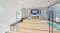 This contemporary fitness center is designed to support a wide range of exercise routines and wellness activities. The main gym floor is expansive and thoughtfully organized, featuring an impressive selection of Hoist-brand strength training equipment, including cable machines, multi-function racks, and a full dumbbell rack. Cardio machines—such as treadmills, ellipticals, and a stair climber—are arranged along the perimeter, complemented by track lighting overhead that keeps the space bright and energized. Large floor-to-ceiling glass walls and windows allow abundant natural light to flood the room and offer views of the surrounding outdoor terrace and cityscape, creating an open, motivating atmosphere.

Adjacent to the main gym is a dedicated studio room, separated by sleek glass partitions. This lighter, airy space features light wood-toned flooring, full-length mirrors, and a wall-mounted flat-screen display, making it well-suited for yoga, stretching, or group fitness classes. A built-in storage cubby provides organized space for equipment such as exercise balls and mats, while a bench near the entrance adds a practical touch. The overall design of the fitness center balances functionality with a clean, modern aesthetic, making it an inviting environment for all fitness levels.
