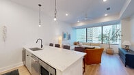 This contemporary urban residence is thoughtfully designed with clean lines, a neutral palette, and high-quality finishes throughout. Light wood flooring flows consistently across the space, connecting each room with a sense of warmth and cohesion.

The open-concept kitchen is a standout feature, equipped with stainless steel appliances, quartz countertops, a large central island with seating, and sleek white cabinetry. Pendant lighting with clear glass shades adds a stylish focal point above the island.

The living area is bright and inviting, anchored by floor-to-ceiling windows that frame impressive city skyline views. A ceiling fan, recessed lighting, and a generous sectional sofa make this an ideal space for both relaxing and entertaining.

The bedroom continues the modern aesthetic with large windows offering sweeping urban views, creating an airy and light-filled retreat. Adjacent is a spacious walk-in closet outfitted with multi-tiered shelving and hanging rods for ample storage.

Two well-appointed bathrooms serve the residence. The primary bath features a double-sink vanity with warm wood cabinetry, a glass-enclosed walk-in shower with tile surround, and illuminated mirrors. The secondary bath is equally refined, with stone-look tile flooring and matte black hardware throughout. A hallway with built-in shelving connects the private areas of the home seamlessly.