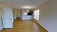 This single-floor space showcases an open-concept design that seamlessly connects the living area to the kitchen. The living space is finished with warm, light-toned hardwood-style flooring that extends throughout, contributing to a bright and cohesive atmosphere. Natural light enters through multiple windows fitted with white blinds, enhancing the airy feel of the interior.

A chandelier with multiple arms serves as a focal point overhead, adding a decorative touch to the dining or living area. The kitchen is defined by a raised breakfast bar counter that provides both a functional workspace and a natural dividing element between the living and cooking areas. Dark wood cabinetry lines the kitchen walls, contrasting with the lighter tones of the rest of the space, while black appliances — including a refrigerator and microwave — complete the kitchen's aesthetic.

A hallway visible toward the rear leads to additional rooms, with white panel doors providing access to what appear to be closets or utility spaces. The neutral wall tones and clean white trim throughout create a versatile, move-in ready environment suitable for a variety of interior styles.