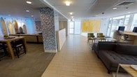 This single-floor space presents a well-designed lobby or reception area with a clean, contemporary aesthetic. The layout is open and spacious, with light-toned large-format tile flooring that extends throughout, creating a seamless and polished look underfoot. A striking floor-to-ceiling mosaic tile accent column serves as a bold architectural centerpiece, featuring a blend of blue, gray, and white tiles that add visual texture and depth to the space.

Adjacent to the column, a reception or service counter finished in white cabinetry provides a functional workspace. To the right, a seating area is furnished with green upholstered chairs and a gray sofa arranged around a small coffee table, offering a comfortable waiting area for visitors. Large windows along the far wall allow ample natural light to filter in, with yellow panel accents adding a vibrant, energetic touch to the atmosphere.

The ceiling is finished with recessed flush-mount lighting that provides consistent, even illumination throughout. The overall ambiance is professional, welcoming, and modern, making this space well-suited for a corporate office, medical facility, or institutional building entrance.