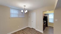 This single-floor residence offers a clean, functional layout designed for comfortable everyday living. The dining area serves as a welcoming central space, finished with warm hardwood-style flooring that extends throughout. A elegant chandelier with brushed nickel accents and frosted glass shades provides focused overhead lighting, while a window fitted with horizontal blinds allows for natural light and privacy control. Neutral, light beige walls create a bright and airy atmosphere, complemented by crisp white trim and baseboards that lend a polished, cohesive look. A white-paneled interior door connects to an adjacent space, adding to the home's clean aesthetic. The kitchen, visible through an open passageway, is well-equipped with dark wood cabinetry and full-sized black appliances, including a refrigerator and a gas range, offering a practical and organized cooking environment. A wall-mounted thermostat and floor vent indicate a climate-controlled interior for year-round comfort. The overall design is straightforward and move-in ready, with consistent finishes throughout that create a unified and inviting living environment.