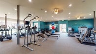 This spacious, single-floor fitness center is designed to support a full range of workout routines in a bright and motivating environment. The interior is defined by bold teal accent walls adorned with inspirational quotes in stylish script lettering, creating an energizing atmosphere throughout the space.

The cardio section is generously outfitted with a row of TRUE-brand treadmills and elliptical machines, all positioned along a wall of large windows that flood the area with natural light and provide views of the surrounding outdoor landscape. A wall-mounted flat-screen TV is conveniently positioned for use during cardio sessions.

The strength training area features multiple selectorized cable and resistance machines placed on protective rubber flooring, alongside adjustable benches and a dumbbell rack. Free-weight and functional training options are further supported by a medicine ball rack and a collection of stability balls in various sizes.

Rowing machines are also available for low-impact, full-body conditioning. Mirrored walls expand the sense of space and allow users to monitor their form. Recessed and ceiling-mounted lighting, along with ceiling fans, ensure the environment remains comfortable.

A dedicated hallway area includes dual water fountains and additional treadmills, leading to further access points within the facility. Carpeted flooring throughout contributes to a clean, polished finish.
