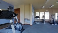 This single-floor fitness room is designed to provide a practical and comfortable workout space. The room features neutral beige walls with dark wood baseboards and is finished with durable, low-pile grey carpeting suited for exercise equipment. A structural column divides the space, creating a natural layout separation between different workout zones. Natural light filters in through a set of large windows on one wall, offering views of surrounding greenery and contributing to an open, airy atmosphere. The room is equipped with several cardio machines, including a stationary bike, a treadmill, and what appears to be an elliptical trainer, providing a range of cardiovascular workout options. A dual-basin water fountain station is conveniently installed along one wall, allowing users to stay hydrated during workouts. A wall-mounted display or monitor is also present near the equipment. Access to the room is provided through a solid wood door. Ceiling-mounted ventilation and lighting fixtures help maintain a comfortable environment throughout the space.