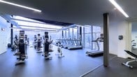 This contemporary fitness center is designed with both function and aesthetics in mind, offering a bright, open workout environment that motivates and inspires. The space is defined by expansive floor-to-ceiling windows that line the perimeter, flooding the interior with natural light and providing sweeping views of the surrounding outdoor landscape — a refreshing backdrop for any workout session.

The cardio zone is a standout feature, housing an extensive row of treadmills, elliptical trainers, stationary bikes, and other cardio equipment, all thoughtfully arranged to take full advantage of the panoramic window views. The smooth, durable flooring in a neutral gray tone provides a clean, professional finish underfoot and complements the sleek, minimalist aesthetic of the space.

Overhead, a dark ceiling with integrated linear lighting fixtures adds a modern industrial touch while ensuring consistent, well-distributed illumination throughout. The combination of natural and artificial lighting creates an energizing ambiance that is both functional and visually appealing.

The layout is spacious and uncluttered, allowing for comfortable movement between equipment. The overall design reflects a high-end, purpose-built fitness facility that prioritizes user experience, natural connectivity, and a polished, modern atmosphere.