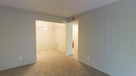 This single-floor residential space offers a clean, functional layout with a warm and neutral aesthetic throughout. The interior is finished with wall-to-wall carpeting in a soft beige tone, creating a cohesive and comfortable feel across the living areas. Walls are painted in light, muted shades that contribute to a calm and airy atmosphere, even in the absence of abundant natural light.

The main living area opens seamlessly into an adjacent dining or secondary room, defined by a partial wall with a wide pass-through opening. This architectural detail creates a sense of spatial flow while maintaining subtle separation between zones. The adjoining room features a pendant light fixture centered overhead, adding a touch of understated elegance and providing warm, focused illumination.

A hallway branching off from the main area leads toward additional rooms, hinting at a well-organized floor plan. Visible HVAC vents in both the ceiling and floor indicate a comprehensive climate control system integrated throughout the space. Electrical outlets are thoughtfully placed along the baseboards, and light switches are conveniently positioned near key transition points.

Overall, the space presents a straightforward, move-in-ready interior with consistent finishes, a neutral palette, and a practical layout well-suited for everyday living.