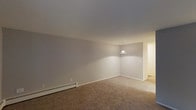 This single-floor residence offers two bedrooms and one and a half bathrooms within a well-proportioned layout. The main living area features wall-to-wall carpeting in a warm neutral tone, contributing to a cozy atmosphere. Smooth, light gray walls provide a clean, understated backdrop throughout the space. A pendant light fixture is positioned at the transition between the living and adjacent areas, providing focused overhead illumination. A baseboard heating unit runs along one wall, ensuring efficient climate control. The open sightlines between the living space and the connecting hallway or adjoining room enhance the overall sense of flow. The neutral color palette of both the walls and flooring creates a versatile environment adaptable to a variety of furnishing styles. The consistent finishes across the visible areas suggest a cohesive design approach throughout the home. With its straightforward single-level layout and practical features, this residence offers a comfortable and easy-to-navigate living environment suited for a range of lifestyles.