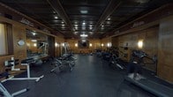 Located at 50 N Illinois St in downtown Indianapolis, this fitness center is a striking blend of historic architectural character and modern exercise amenities. The space is defined by its warm, richly stained wood-paneled walls featuring geometric raised-panel detailing, complemented by deep coffered ceilings with dark wood beams that lend a sense of grandeur and heritage to the room. A decorative frieze running along the upper walls depicts historic train motifs, nodding to the building's storied past. Ambient wall sconces and modern pendant lighting — including a sleek circular ring light fixture — provide layered illumination throughout the space. The rubber-floored fitness area is well-equipped with a comprehensive selection of cardio machines, including treadmills, ellipticals, stationary bikes, and a stair climber, as well as strength training equipment such as a cable machine, adjustable benches, and a dumbbell rack. A large wall mirror on one side expands the sense of space and supports functional training. A flat-screen television is mounted on the wall for entertainment during workouts. A glass-partitioned reception or attendant area is visible near the entrance, alongside large glass doors that allow natural light to filter in. The overall ambiance is uniquely distinguished — a fitness facility that retains the elegance and craftsmanship of its historic surroundings.