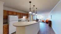 This single-floor residence offers a thoughtfully designed layout that combines comfort and functionality. The open-concept kitchen and living area serves as the heart of the unit, anchored by a curved white breakfast bar island with elegant pendant lighting featuring ornate bronze fixtures. The kitchen is equipped with warm honey-toned wood cabinetry, a white range, and a full-size refrigerator, with crown molding adding a refined architectural touch. The kitchen flows seamlessly into the spacious living room, which features durable hardwood-style flooring, abundant natural light from large windows, and a cozy reading nook with an oversized window framing outdoor views. The bedroom is a comfortable retreat with plush carpeted floors, dual windows that invite natural light, and generous closet space. The primary bathroom includes a mirrored closet, a vanity with wood cabinetry, and a separate water closet for added privacy. A second full bathroom features a wood vanity, a shower-tub combination, and hardwood-style flooring. Throughout the unit, neutral wall tones, crown molding in key areas, and consistent warm-toned flooring create a cohesive and inviting atmosphere well-suited for everyday living.