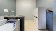 This single-floor shared laundry facility is designed for practical, efficient use. The space is divided into two connected areas, each painted in calming blue-grey tones and finished with durable light-colored tile flooring throughout.

The main washing area features a row of top-loading washing machines, providing ample capacity for multiple users simultaneously. A wall-mounted air conditioning unit ensures a comfortable environment year-round. Natural light filters in through a window, brightening the space, while a small folding table and chair offer a convenient spot for waiting or sorting laundry.

The adjacent drying area is equipped with several stacked dryer units, maximizing the use of vertical space while keeping the floor area open and easy to navigate. A dedicated folding counter with a dark base provides a sturdy workspace for handling clean laundry. An electrical panel is accessible on the wall, and informational notices are posted for user reference. A clearly marked emergency call station is also present near the exit door, reflecting attention to occupant safety.

Overall, the facility is clean, well-organized, and functionally laid out, offering residents a straightforward and comfortable laundry experience.