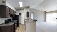 This single-story unit is designed with a practical, open layout that connects key living areas cohesively. The living room serves as a focal point, showcasing vaulted ceilings with a textured finish, a ceiling fan, and a fireplace framed by a clean white mantel. Natural light enters through a sliding glass door that leads to an outdoor area, enhancing the sense of openness. The carpeted floors carry through the living and bedroom spaces, contributing to a comfortable, neutral aesthetic throughout.

The kitchen is efficiently arranged in a galley-style configuration, featuring dark espresso cabinetry paired with granite-style countertops. Black appliances, including a range and refrigerator, are integrated alongside a double stainless steel sink. A peninsula with additional counter space connects the kitchen to the living area, offering added functionality. Wood-look flooring runs through the kitchen, providing a durable, easy-to-maintain surface. A louvered closet door near the kitchen provides convenient additional storage.

The bedrooms are generously proportioned, each finished with soft carpeting, neutral gray walls, and windows that allow natural light. The bathroom includes dark cabinetry with matching granite-style countertops, wood-look flooring, and a full tub-shower combination with white tile surround.