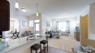 This single-floor residence showcases a thoughtfully designed open-concept layout that seamlessly integrates the kitchen, dining, and living areas into one welcoming, cohesive space. The kitchen features a granite-topped breakfast bar with stylish upholstered counter stools, an undermount sink, and pendant lighting that adds a contemporary accent overhead. Rich hardwood-style flooring anchors the kitchen and dining zone, while plush carpeting transitions naturally into the spacious living area, adding warmth and comfort underfoot. The living room is generously proportioned, bathed in natural light streaming through large windows dressed with elegant blue grayish curtains. Vaulted ceilings lend an airy, expansive quality to the space, enhancing the sense of openness throughout. The neutral, light-toned walls provide a clean and versatile backdrop, complementing the classic yet casual furnishings. A dedicated entry area near the front door includes a small accent table, adding a functional and welcoming touch to the arrival experience. Throughout the space, the interplay of natural light, open sightlines, and carefully chosen finishes creates a bright and inviting atmosphere that feels both comfortable and well-appointed.