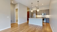 This thoughtfully designed single-floor residence showcases a clean, contemporary aesthetic throughout. The open-concept layout connects the kitchen and living area seamlessly, anchored by warm-toned hardwood-style flooring that runs throughout the main living spaces. The kitchen is a standout feature, equipped with stainless steel appliances including a gas range, built-in microwave, and French door refrigerator. Sleek gray flat-front cabinetry with matte black hardware pairs with crisp white countertops and a classic subway tile backsplash. A central kitchen island with an integrated sink and globe pendant lighting above adds both function and style to the space. The open living area offers generous room to arrange furnishings, with neutral gray walls providing a versatile backdrop. The bedroom is carpeted for comfort and benefits from natural light through a pair of large windows that overlook a private balcony, with views of the surrounding neighborhood. The bathroom features a soaking tub with a tiled surround in a clean white stacked-tile pattern, chrome fixtures, and stone-look tile flooring. A dedicated laundry area houses a stacked front-load washer and dryer alongside wire shelving for additional storage, completing this well-appointed residence.
