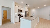 This single-floor residence is designed with a clean, contemporary aesthetic that emphasizes bright, open living. The kitchen serves as a central highlight, featuring a large island with a granite countertop and an integrated sink, tall white shaker-style cabinetry, a white subway tile backsplash, and a full suite of stainless steel appliances including a refrigerator, range, and microwave. Recessed lighting illuminates the space efficiently. Light wood-look flooring runs consistently throughout the unit, lending a cohesive and airy feel to every room.

The living area is generously sized and filled with natural light from multiple windows and a glass door that opens to the outdoors, with a ceiling fan overhead. The bedrooms are bright and neutral, each with windows fitted with blinds to control light levels.

The bathrooms are finished to match the kitchen's modern standard, featuring granite vanity countertops, white cabinetry with brushed chrome hardware, and large mirrors. One bathroom includes a glass-enclosed walk-in shower and built-in open shelving for added storage. Throughout, the design prioritizes clean lines, neutral tones, and functional finishes.