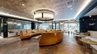 Located at 5880 Inwood Dr in Houston's Galleria area, this expansive single-floor amenity lounge offers a sophisticated, multi-zone gathering space thoughtfully designed with high-end finishes and a cohesive yet varied aesthetic throughout. The main lounge area immediately draws attention with rich herringbone-patterned hardwood flooring, a striking textured concrete ceiling, and a dramatic circular drum pendant light that anchors a curved, caramel-toned leather sectional sofa arrangement — an inviting centerpiece ideal for social gatherings. Large windows along the perimeter flood the space with natural light and offer views to the outdoors. Transitioning deeper into the space, the design shifts to a bold chevron-patterned carpet beneath exposed natural wood ceiling beams, creating a warmer, more casual atmosphere. Sculptural concrete archways frame intimate booth-style seating nooks, perfect for smaller conversations or focused gatherings. A foosball table and lounge seating with a mounted flat-screen television offer additional recreational options. A communal bar area features a substantial reclaimed wood counter, bar-height seating, and a row of brushed brass pendant lights suspended from the concrete ceiling, complemented by dark brick accent walls and large-scale artwork. Sleek black steel-framed glass partitions and doors unify the various zones while maintaining an open, connected feel throughout.