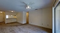 This single-floor unit offers two bedrooms and two bathrooms within a well-utilized floor plan. The living area is spacious enough to accommodate a variety of furniture arrangements and features wall-to-wall carpeting that adds warmth underfoot. A ceiling fan with an integrated light fixture is centrally positioned overhead, providing both air circulation and ambient lighting to the main living space.

Neutral, light-toned walls throughout the unit create a clean, versatile backdrop suitable for a range of décor styles. A partial wall divides the living area, creating a subtle sense of separation while maintaining an open feel between spaces. A hallway leads to the bedrooms, offering a degree of privacy from the main living and dining areas.

Adjacent to the living space, a pass-through or bar-height counter connects to the kitchen area, visible in the background, which benefits from its own lighting. The overall layout prioritizes functionality, with straightforward traffic flow between rooms. The unit's single-floor design ensures easy accessibility throughout, making it a practical and comfortable living space.