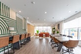 Located at 100 Manett Crescent in Brampton, Ontario, this thoughtfully designed single-floor amenity space offers a well-appointed layout across four distinct areas. The open-concept lounge is a standout feature, bathed in natural light streaming through large floor-to-ceiling windows fitted with roller shades. Warm cognac leather sofas, rattan accent chairs, and lush indoor plants create an inviting, contemporary atmosphere complemented by light hardwood flooring throughout.

A slatted wood partition elegantly separates the lounge zones, adding architectural depth while maintaining an open, airy feel. Recessed lighting and crisp white ceilings contribute to the clean, modern aesthetic. A dedicated seating nook with upholstered chairs and a side table provides a more intimate setting within the lounge area.

The conference room accommodates a full boardroom-style table with ergonomic seating, framed by expansive windows overlooking greenery — ideal for productive meetings in a bright, professional environment.

The fully equipped kitchen features slate-blue cabinetry, white quartz countertops, stainless steel appliances including a range with hood vent and refrigerator, and a central island with bar seating. A bold decorative mural adds a creative accent, rounding out a space that balances function with refined, modern style.