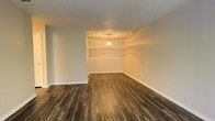 This single-floor space presents a well-designed interior that balances functional simplicity with thoughtful architectural details. The main living area immediately draws the eye with its rich, dark hardwood-style flooring, which runs throughout and provides a warm, grounding contrast to the neutral, light-toned walls. A standout feature of the space is the defined dining or sitting nook, distinguished by a tray ceiling with a decorative circular medallion and a centered pendant light that casts a soft, inviting glow. Built-in shelving flanks the nook area, offering both display and storage opportunities while adding architectural character to the room. The textured ceiling throughout the space adds subtle visual depth, complementing the overall clean aesthetic. The open layout allows for flexible furniture arrangement, making the space adaptable to a variety of uses. Smooth, painted walls in a soft neutral tone keep the environment bright and airy, while the transitions between areas feel natural and unobstructed. Overall, this space offers a cohesive and practical interior with distinctive design touches that elevate it beyond the ordinary.