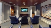 This private movie theater is a thoughtfully designed single-floor entertainment space that blends comfort with a sophisticated aesthetic. The main viewing area is anchored by a wall-mounted flat-screen television, positioned above a low wood-slatted media console and flanked by doorways that lend a sense of depth and accessibility to the room. Seating is arranged for optimal viewing, with multiple plush navy blue leather recliners and ottomans oriented toward the screen, creating an intimate and inviting atmosphere. Warm wood panel ceilings with recessed lighting add a rich, refined character to the space, while the neutral carpeted floor ties the room together. Floor-to-ceiling gray curtain panels provide an option to control ambient light and enhance the cinematic experience. Adjacent to the main viewing area is a casual lounge corner, featuring a round wooden high-top table with coordinating blue wire-frame bar stools — ideal for socializing before or after a screening. A large decorative Scrabble board mounted on the wall adds a playful, recreational touch. Wall sconces provide soft accent lighting throughout, complementing the warm tones of the wood paneling. A security camera and exit signage are also present, underscoring the space's practical and safety-conscious design. Overall, the space offers a versatile, comfortable environment for both movie viewing and casual gathering.