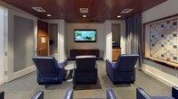 This private movie theater is a thoughtfully designed single-floor entertainment space that blends comfort with a sophisticated aesthetic. The main viewing area is anchored by a wall-mounted flat-screen television, positioned above a low wood-slatted media console and flanked by doorways that lend a sense of depth and accessibility to the room. Seating is arranged for optimal viewing, with multiple plush navy blue leather recliners and ottomans oriented toward the screen, creating an intimate and inviting atmosphere. Warm wood panel ceilings with recessed lighting add a rich, refined character to the space, while the neutral carpeted floor ties the room together. Floor-to-ceiling gray curtain panels provide an option to control ambient light and enhance the cinematic experience. Adjacent to the main viewing area is a casual lounge corner, featuring a round wooden high-top table with coordinating blue wire-frame bar stools — ideal for socializing before or after a screening. A large decorative Scrabble board mounted on the wall adds a playful, recreational touch. Wall sconces provide soft accent lighting throughout, complementing the warm tones of the wood paneling. A security camera and exit signage are also present, underscoring the space's practical and safety-conscious design. Overall, the space offers a versatile, comfortable environment for both movie viewing and casual gathering.