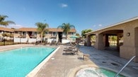 This outdoor amenity space is centered around a large, clear-blue swimming pool that serves as the focal point of the area. The pool is complemented by an adjacent hot tub/spa, complete with stainless steel entry rails for easy access. Notably, an ADA-compliant pool lift is installed poolside, ensuring accessibility for all users. The expansive concrete pool deck offers ample room for relaxation and is furnished with multiple reclining lounge chairs and a shaded seating area featuring a green market umbrella and additional chairs — ideal for outdoor gatherings. A generously sized covered patio with a stucco-finished exterior and sturdy support columns provides a shaded retreat adjacent to the pool area. The patio is accessed through a set of white double French doors with slatted blinds. A water fountain and utility area with an emergency shut-off switch are also integrated into the facility's infrastructure. The surrounding landscape is adorned with mature palm trees and lush greenery, contributing to a warm, resort-like atmosphere. The multi-story residential buildings visible in the background, finished in neutral earth tones with balconies and a wrought iron perimeter fence, frame the space and reinforce the sense of a secure, community-oriented outdoor environment.