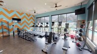 This fitness center offers a well-rounded workout environment designed with both functionality and visual appeal in mind. The space is defined by a wall of large, floor-to-ceiling windows along one side, allowing abundant natural light to fill the room while providing views of an outdoor pool and landscaped surroundings. A bold orange and teal chevron accent wall serves as a striking focal point, complementing the aqua-toned walls throughout.

The cardio zone lines the window wall and features multiple treadmills and elliptical machines, each equipped with individual monitors. Ceiling fans are positioned throughout to maintain comfortable airflow during workouts. The strength training area occupies the opposite side of the room and includes cable machines, a multi-functional press station, adjustable benches, and a well-stocked rack of kettlebells and free weights in varying sizes.

Large interior glass partitions visually expand the space and enhance the open, airy feel of the room. Dark hardwood-style flooring runs throughout, providing a polished, durable surface. A glass door leads directly to an outdoor patio area, offering convenient access to exterior amenities. The overall design balances an energetic aesthetic with practical, comprehensive fitness offerings.