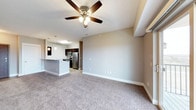 This single-floor residence showcases an open-concept layout that connects the living area to the kitchen in a seamless, functional flow. The living area is finished with plush wall-to-wall carpeting in a warm neutral tone, offering a comfortable and inviting atmosphere. A ceiling fan with an integrated light fixture serves as a practical focal point overhead, contributing to both air circulation and ambient lighting.

The kitchen is positioned at the far end of the space and features dark cabinetry paired with stainless steel appliances, including a refrigerator, range, and microwave. A peninsula counter with pendant lighting overhead adds a contemporary touch while providing additional prep and seating space. The kitchen area transitions smoothly from the carpeted living space via hard-surface flooring, creating a clear and practical delineation between the two zones.

The walls throughout are finished in a soft, neutral greige tone that complements the overall color palette, while white baseboards provide clean contrast. The open design allows light to travel freely across the space, enhancing the sense of openness. A wall-mounted unit is also visible, offering supplemental climate control options.
