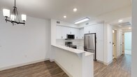 This single-floor space showcases a clean, modern aesthetic defined by bright whites, warm wood-tone flooring, and thoughtful design details throughout. The open-concept kitchen is a standout feature, outfitted with crisp white shaker-style cabinetry that extends to the ceiling, providing generous storage. Stainless steel appliances — including a French door refrigerator, electric range, and built-in microwave — lend a polished, contemporary feel. A peninsula with an extended white countertop serves as both a prep surface and a casual breakfast bar, seamlessly connecting the kitchen to the adjacent living area. Recessed lighting and a sleek flush-mount fixture illuminate the space evenly, enhancing the bright, airy atmosphere created by the all-white walls and ceilings. The rich, dark hardwood-style flooring grounds the space and adds warmth, creating a striking contrast against the light cabinetry and countertops. The layout is efficient yet open, making excellent use of the available footprint. Overall, the space presents a fresh, move-in-ready environment with a cohesive design language that balances functionality with modern style.
