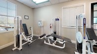 This fitness center offers a comprehensive workout environment spread across a single-floor layout designed to accommodate a wide range of exercise needs. The space is finished with durable rubber flooring throughout, providing both safety and functionality underfoot. Neutral-toned walls and a drop ceiling with recessed fluorescent lighting create a clean, practical atmosphere, while ceiling fans help maintain airflow and comfort during workouts.

The main area features an array of cardio equipment, including treadmills, elliptical machines, and a stationary bike, all positioned near windows fitted with blinds that allow natural light to filter in. A wall-mounted television adds entertainment for those logging time on cardio machines. A beverage and snack vending machine is conveniently located near the entrance for post-workout refreshment.

A dedicated strength training section houses multiple cable and weight machines, adjustable benches, and a rack of free weights, catering to both beginners and experienced lifters. Full-length mirrors line one wall, enhancing the sense of space while allowing users to monitor their form. A separate room provides additional strength machines and a fire safety station, ensuring a well-organized and safety-conscious environment. Framed certifications and informational displays are posted throughout, adding a professional touch to the overall facility.