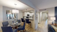 This single-floor residence offers a thoughtfully designed layout that balances comfort and functionality. The living room serves as the heart of the home, featuring light wood-style flooring, a bold navy accent wall adorned with large-scale artwork, and a sliding glass door that opens to a private balcony, filling the space with natural light and outdoor views. Adjacent to the living room, the dining area accommodates a round glass-top table beneath a contemporary drum pendant light, creating an inviting setting for meals and gatherings. The kitchen is well-equipped with white shaker cabinetry, granite countertops, a classic subway tile backsplash, stainless steel appliances, and a breakfast bar with seating — seamlessly connecting to the dining space. A hallway leads to two generously sized bedrooms, each featuring wood-style flooring, ceiling fans, and ample closet space. The primary bedroom includes a walk-in closet for added storage convenience. Both bathrooms are finished with granite-topped white vanities and tub-shower combinations, offering a clean and polished aesthetic. Throughout the residence, neutral tones are accented by stylish décor elements, creating a cohesive and welcoming ambiance.