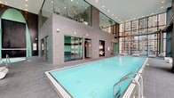 This upscale amenity floor offers a comprehensive wellness experience set against a striking urban backdrop. The centerpiece is a generously sized indoor lap pool, enclosed by soaring floor-to-ceiling glass walls that flood the space with natural light and frame panoramic city skyline views. Sleek grey tile flooring and patterned stone wall cladding lend the pool area a refined, contemporary aesthetic, while recessed lighting enhances the atmosphere during evening hours.

Adjacent to the pool, a dedicated hot tub alcove features mosaic tile surrounds in deep black and teal tones, complemented by a striking water wall feature and large windows overlooking the cityscape below. A spacious open-air shower room finished entirely in vibrant aqua-green mosaic tiles provides a luxurious rinse experience, with high-end chrome fixtures and views to the exterior.

The sunroom lounge area is a standout feature, wrapped in dramatic floor-to-ceiling glass on multiple sides and furnished with comfortable wicker chaise lounges. This serene retreat connects directly to an expansive outdoor terrace, where additional lounge seating is arranged to take full advantage of the sweeping urban views. A mezzanine-level viewing area overlooks the pool below, completing this thoughtfully designed, resort-style wellness destination in the heart of the city.