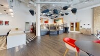 This thoughtfully designed single-floor commercial space blends creativity and functionality across several distinct zones. The main gathering area makes an immediate impression with its soaring exposed-ceiling design, showcasing an eye-catching installation of sculptural acoustic panels suspended overhead alongside pendant lighting. A central multi-level lounge structure with built-in bunk-style seating and integrated staircases serves as a striking focal point, surrounded by open hardwood flooring and vibrant accent furniture in red and neutral tones. Bold, colorful artwork adorns the walls, adding an energetic personality to the space. A separate lounge area features floor-to-ceiling frosted glass windows that flood the room with diffused natural light, complemented by an intricate textured accent wall, an embedded flat-screen display, and a curated mix of white and red upholstered seating beneath dramatic wire-frame pendant chandeliers. A quieter nook offers a long upholstered bench seat set against a chevron-patterned wallcovering, paired with contemporary pod-style chairs and round tables — ideal for casual meetings or relaxation. A compact kitchenette area rounds out the space, finished with light wood cabinetry, clean white countertops, patterned flooring, and pop-art framed prints. Throughout, the space balances bold design choices with practical amenities, creating a dynamic and welcoming environment.