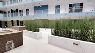This multi-story building is centered around a striking open-air courtyard that serves as its architectural focal point. The courtyard is framed by the building's upper floors, which feature white-railed balconies and a clean, contemporary facade finished in soft blue and white tones. Abundant natural light floods the space from above, creating a bright and airy atmosphere throughout the day.

The courtyard itself is thoughtfully landscaped with large, white rectangular planters filled with tall ornamental grasses that rise dramatically, adding a strong vertical design element and a sense of natural privacy. The light-toned, smooth flooring runs throughout the open space, lending a clean and cohesive aesthetic.

A standout feature of the courtyard is a centrally positioned water feature clad in rich brown tile, complete with a cascading water element that introduces a soothing, ambient quality to the environment. Concrete seating elements are integrated into the layout, providing functional gathering areas within the courtyard.

Ground-floor units are directly accessible from the courtyard via sage-green painted doors, blending seamlessly with the overall color palette. Security and wayfinding elements are thoughtfully incorporated throughout. The design balances architectural structure with natural elements, creating a welcoming and visually cohesive communal space.