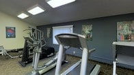 This fitness room is a dedicated exercise space designed with functionality and practicality in mind. The room features a cool-toned, slate-blue color scheme on the walls, lending a focused and energizing atmosphere ideal for workouts. Overhead skylights and recessed flush lighting ensure the space is well-lit throughout the day, creating a bright and inviting environment.

The room is equipped with a selection of cardio machines, including treadmills and an elliptical trainer from reputable brands such as True Fitness, offering users quality equipment for cardiovascular exercise. The machines are thoughtfully arranged to maximize use of the available floor space, with protective rubber mats placed beneath the equipment for safety and floor preservation.

A wall-mounted mirror is featured on one side, a common and practical addition to fitness spaces that allows users to monitor their form during exercise. Motivational and instructional posters are displayed on the walls, adding an informative and encouraging element to the room's design. A window with vertical blinds provides a source of natural light and ventilation. The overall layout and design of this fitness room make it a convenient and functional amenity well-suited for regular use.