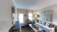 This well-designed single-floor residence exudes a refined and welcoming ambiance, blending contemporary style with classic touches throughout. The living area serves as the heart of the space, showcasing a sophisticated neutral color palette of warm grays and taupe tones that lend a sense of calm and cohesion. Crown molding lines the ceiling, adding an elegant architectural detail that elevates the overall aesthetic.

Natural light pours in through multiple windows dressed with white blinds and soft draping curtains, brightening the room and creating an airy atmosphere. The living area is thoughtfully furnished with a plush gray sofa accented by decorative pillows, a rich brown leather armchair, and a sleek metallic coffee table adorned with tasteful greenery. A dark console unit lines one wall, styled with candle holders and decorative vases, contributing to the room's layered and curated look.

Statement décor pieces, including a sunburst mirror and an eye-catching sunburst wall accent, introduce golden tones that add warmth and visual interest. A framed nature-inspired artwork completes the wall arrangement, tying together the organic and contemporary elements of the design. Dark hardwood-style flooring grounds the space with depth and contrast, while a richly patterned area rug defines the seating arrangement and adds texture. Overall, the space offers a polished and livable environment.