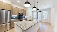 This well-designed 1-bedroom, 1-bathroom unit offers a thoughtful layout that maximizes its compact footprint. The open-concept kitchen and living area serves as the heart of the space, anchored by a large granite-topped island with an undermount sink and stylish pendant lighting overhead. The kitchen is equipped with stainless steel appliances, including a range, microwave, and refrigerator, complemented by warm wood cabinetry and a sleek subway tile backsplash. Light hardwood flooring flows throughout the main living areas, lending a clean, contemporary feel.

The living area receives abundant natural light through multiple windows fitted with horizontal blinds, creating a bright and airy atmosphere. A hallway connects the main living space to the private bedroom, which features soft carpeting, a ceiling fan, and generous window coverage for plenty of natural light. The bathroom is well-appointed with a granite-top vanity, wood-toned cabinetry, an illuminated mirror, and a glass-enclosed walk-in shower with marble-style tile surround. A dedicated walk-in closet with built-in shelving is conveniently accessible directly from the bathroom. The overall design blends modern finishes with functional living in a cohesive, neutral palette.