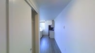 This single-floor space is designed with a straightforward, functional layout that maximizes efficiency. A hallway with smooth white walls and grey wood-look flooring leads toward the kitchen, creating a clean and uncluttered passage through the unit. A sliding door along the hallway provides access to a closet or adjoining room, adding practical storage or flexible use of space.

The kitchen sits at the end of the corridor and benefits from a window that allows natural light to fill the area, brightening the otherwise minimal palette of white cabinetry and dark countertops. The cabinetry is arranged along both walls, offering ample storage, while stainless steel appliances — including a refrigerator and a range — provide a modern touch. The dark backsplash behind the cooking area contrasts neatly with the white upper cabinets, adding visual interest.

Throughout the space, the consistent grey wood-look flooring and crisp white walls contribute to a cohesive, contemporary aesthetic. The overall ambiance is clean, bright, and practical, suited for efficient everyday living.