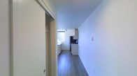 This single-floor space is designed with a straightforward, functional layout that maximizes efficiency. A hallway with smooth white walls and grey wood-look flooring leads toward the kitchen, creating a clean and uncluttered passage through the unit. A sliding door along the hallway provides access to a closet or adjoining room, adding practical storage or flexible use of space.

The kitchen sits at the end of the corridor and benefits from a window that allows natural light to fill the area, brightening the otherwise minimal palette of white cabinetry and dark countertops. The cabinetry is arranged along both walls, offering ample storage, while stainless steel appliances — including a refrigerator and a range — provide a modern touch. The dark backsplash behind the cooking area contrasts neatly with the white upper cabinets, adding visual interest.

Throughout the space, the consistent grey wood-look flooring and crisp white walls contribute to a cohesive, contemporary aesthetic. The overall ambiance is clean, bright, and practical, suited for efficient everyday living.