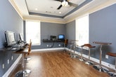 This single-room business center offers a thoughtfully designed workspace finished with warm hardwood flooring and cool slate-blue walls accented by a contrasting cream-toned tray ceiling with recessed lighting. The tray ceiling adds architectural depth and a sense of elegance to the functional space. Multiple wall-mounted workstation shelves with dark granite-style surfaces are equipped with desktop computers and a printer, providing convenient access for users. High-top round tables paired with modern chrome and wood bar-height stools offer additional seating and collaborative workspace areas. Natural light flows generously through several windows fitted with white horizontal blinds, illuminating the room and offering views of the surrounding greenery. A small refreshment station near the entrance features a coffee maker and supplies, adding a welcoming touch. A doorway connects the space to an adjacent room, enhancing accessibility and flow. White trim and six-panel doors throughout contribute to a clean, polished aesthetic. The overall ambiance is professional yet inviting, making this business center a well-equipped and comfortable environment for work and productivity.