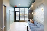 Located in Salem, MA, this conference center offers a thoughtfully designed single-floor layout comprising distinct functional areas that balance professionalism with comfort. The entrance lounge sets an inviting tone with light hardwood flooring, sage-toned walls, and a bold botanical-patterned accent wall adorned with elegant brass wall sconces. A plush sectional sofa in steel blue with warm accent pillows provides a comfortable reception area, complemented by round pedestal side tables and floor-to-ceiling windows that frame views of the surrounding streetscape and fill the space with natural light. Transitioning deeper into the center, a dedicated conference room offers a spacious light wood table surrounded by upholstered swivel chairs, patterned carpet flooring, and soft neutral walls. A wall-mounted display screen supports presentations and meetings, while a striking geometric brass pendant light fixture serves as a focal point overhead. Large windows continue to bring in abundant daylight and outdoor views. A framed abstract artwork adds a refined artistic touch to the room. Together, the spaces create a polished yet welcoming environment well-suited for professional gatherings, collaborative meetings, and events in the heart of Salem.