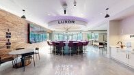 Located at 340 S Market Street in downtown Wilmington, Delaware, the Luxor amenity space is a single-floor community hub designed with a bold, contemporary aesthetic. Polished concrete floors run throughout, lending an industrial-chic foundation to the vibrant interiors.

At the heart of the space is the Luxor bar — a striking circular counter with purple ambient lighting beneath a tiered ceiling medallion, surrounded by plush jewel-toned barstools. The "Love. Life. Here." motto is etched into the floor, reinforcing the community-focused atmosphere.

A dedicated lounge area features generous sectional seating, a sleek linear fireplace set into a stone-clad wall, a large wall-mounted TV, and an eye-catching sculptural chandelier. Expansive windows flood the area with natural light and provide views of the surrounding cityscape.

The game room offers a full-size billiards table, shuffleboard, a wall-length bar-height counter with seating, and a hand-lettered mural reading "don't quit your day dream," adding a spirited, artistic touch.

Additional zones include an "Imbibe" dining nook framed by a richly textured reclaimed wood accent wall and a multi-screen TV display, a kitchenette for resident use, and intimate seating vignettes adorned with original artwork and live-edge wood mirrors. Decorative laser-cut screens provide elegant spatial separation throughout.