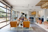 This thoughtfully designed single-floor amenity lounge showcases a modern yet warm aesthetic, blending industrial and contemporary elements throughout its expansive open-concept layout. Large-format slate tile flooring flows continuously across the space, anchoring each distinct zone while maintaining visual cohesion.

The main entry welcomes guests with generous ceiling height, floor-to-ceiling glass doors framed in dark metal, and abundant natural light that spills across the polished tile. Adjacent to the entry, full-height black-framed windows and retractable glass garage doors dissolve the boundary between the interior lounge and an outdoor patio area, offering seamless indoor-outdoor connectivity.

Multiple seating arrangements are distributed throughout, including tan leather lounge chairs with wood frames, a velvet green sofa beneath a striking slatted wood ceiling feature, and curated communal dining tables with mixed seating. Decorative pendant lighting—ranging from oversized matte black fixtures to ornate gold woven pendants—adds dimension and warmth overhead.

A built-in wet bar with navy cabinetry, light quartz countertops, a marble backsplash, and under-counter refrigeration provides a functional hospitality station. Curated artwork, organic sculptural accents, and lush potted greenery complete the refined, welcoming ambiance.