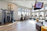 This expansive fitness studio is designed to support a wide range of workout styles and fitness goals. The space is organized into several distinct zones, all connected by light wood-tone flooring and bright, white walls that create a clean and energizing atmosphere. Soaring ceilings and an abundance of large, black-framed windows flood the interior with natural light while offering open views of the surrounding skyline and cityscape.

The cardio zone is well-appointed with a row of Matrix treadmills, elliptical trainers, stair climbers, and stationary cycling bikes, all positioned to take advantage of the panoramic window views. A ceiling-mounted flat-screen TV provides entertainment throughout the space.

The strength and functional training area features a cable cross machine, a heavy punching bag, TRX suspension trainers, and a fully stocked equipment rack holding kettlebells, medicine balls, stability balls, resistance bands, and yoga mats. A ballet barre along one wall adds versatility for stretching and low-impact training. Additional free weights and a weight bench round out the strength offerings.

A ceiling fan adds comfort, and the studio is accessed via a key-fob secured entrance. The overall ambiance is modern, bright, and professional, making this fitness studio a comprehensive facility suited for all fitness levels.