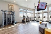 This expansive fitness studio is designed to support a wide range of workout styles and fitness goals. The space is organized into several distinct zones, all connected by light wood-tone flooring and bright, white walls that create a clean and energizing atmosphere. Soaring ceilings and an abundance of large, black-framed windows flood the interior with natural light while offering open views of the surrounding skyline and cityscape.

The cardio zone is well-appointed with a row of Matrix treadmills, elliptical trainers, stair climbers, and stationary cycling bikes, all positioned to take advantage of the panoramic window views. A ceiling-mounted flat-screen TV provides entertainment throughout the space.

The strength and functional training area features a cable cross machine, a heavy punching bag, TRX suspension trainers, and a fully stocked equipment rack holding kettlebells, medicine balls, stability balls, resistance bands, and yoga mats. A ballet barre along one wall adds versatility for stretching and low-impact training. Additional free weights and a weight bench round out the strength offerings.

A ceiling fan adds comfort, and the studio is accessed via a key-fob secured entrance. The overall ambiance is modern, bright, and professional, making this fitness studio a comprehensive facility suited for all fitness levels.